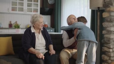 Küçük çocuk büyükannesini ve büyükbabasını ziyaret eder, bayramını ellerini öperek kutlar. Mutlu yaşlı çift koltukta oturur ve küçük torunlarını öper. Torunuyla mutlu bir ailenin portresi.
