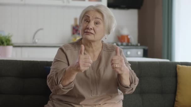 一位脾气暴躁的老妇人坐在沙发上 在照相时竖起大拇指 一位头发灰白 穿着休闲服 疑神疑鬼 满脸不悦的老年妇女 消极的人 — 图库视频影像