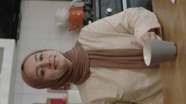 Young woman in hijab works remotely on a tablet computer. The woman drinks coffee or tea and eats roasted hazelnuts while working on the computer in her home kitchen. Freelancer concept.Vertical video