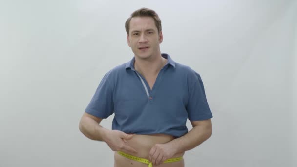 男人用带子测量臀部和腹部 年轻人用带子测量腰围 与白色背景隔离 因为体重增加而不快乐 超重了 肥胖问题 — 图库视频影像