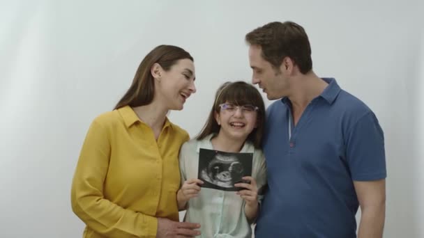 年轻的父母和他们的女儿们看着超声胶卷接近他们未出生婴儿的最后一天 用白色背景为她未出生的妹妹做超声波检查的女孩 理想而快乐的家庭肖像 — 图库视频影像