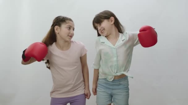 白を背景に二人の少女が並んで立っている カジュアルなTシャツとジーンズを着た精力的な女の子は 彼らがボクシンググローブとどのように強い示しています チームワークの概念 — ストック動画