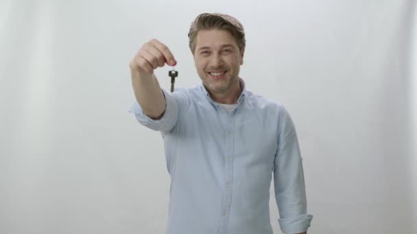 白い背景の前に立つ男は 新しい家の鍵を見せ 幸せそうにカメラを見て笑っている 鍵を持ってカメラを見て笑っている男の肖像 — ストック動画