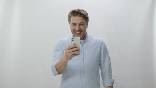 男は白い背景に電話でビデオ通話をする 顔に笑顔で青いシャツの幸せな男は カメラを見て 手を振って 愛する人とビデオ通話を行う オンラインビデオ通話で話す男の肖像 — ストック動画