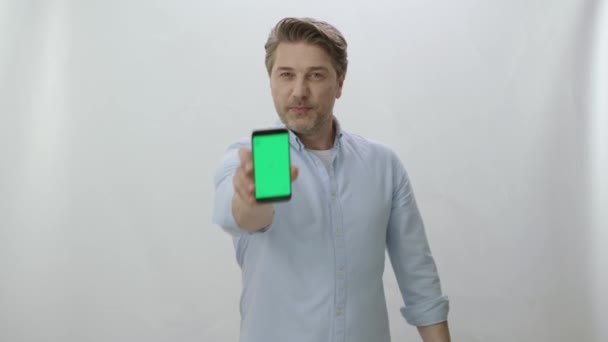 一个年轻可爱的男人站在一个与世隔绝的背景上 展示着一个绿色屏幕的智能手机 一个拿着绿色屏幕的智能手机的有趣男人 — 图库视频影像