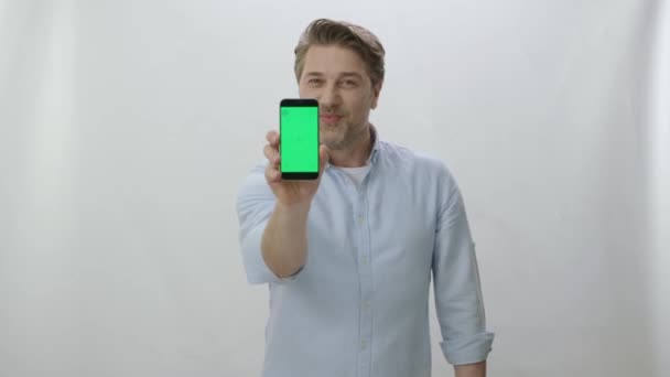 一个年轻可爱的男人站在一个与世隔绝的背景上 展示着一个绿色屏幕的智能手机 一个拿着绿色屏幕的智能手机的有趣男人 — 图库视频影像