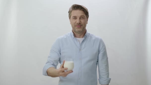 白いスタジオの背景に隔離された笑顔の白人男性 牛乳やバターミルクのカメラと飲料ガラスを見てカジュアルなスーツの若い男 ガラスの中の飲み物は非常に良いことを示す男 — ストック動画