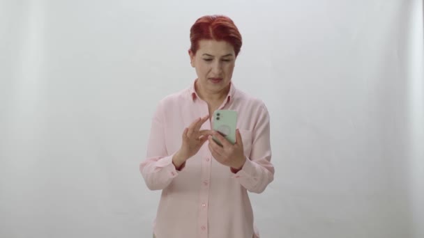 赤い髪の女性は白い背景にスマートフォンでインターネットに時間を費やしています ニュースやテキストメッセージが彼女の携帯電話に到着すると 女性は驚きと非常に怒っています 悪い知らせを読む — ストック動画