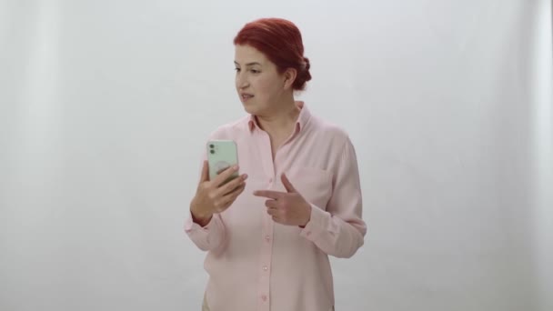 女性は白い背景にスマートフォンを見て 画面の左側に誰かと話をします 質問をします 答えに驚いています あなたは女性が話している任意のメッセージや製品を置くことができます — ストック動画