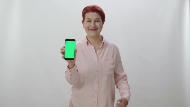 白い背景に緑の画面のスマートフォンを示す赤毛の女性 緑の画面を持つスマートフォンで指で指すゲーマーの女性 — ストック動画