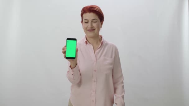 白い背景に緑の画面のスマートフォンを示す赤毛の女性 指で親指を表示し 緑の画面でスマートフォンを指してゲーマーの女性 — ストック動画