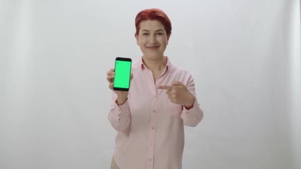 白い背景に緑の画面のスマートフォンを示す赤毛の女性 緑の画面を持つスマートフォンで指で指すゲーマーの女性 — ストック動画