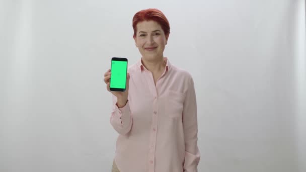 白い背景に緑の画面のスマートフォンを示す赤毛の女性 緑の画面のスマートフォンを指してゲーマーの女性は 勝利のサインで指 — ストック動画