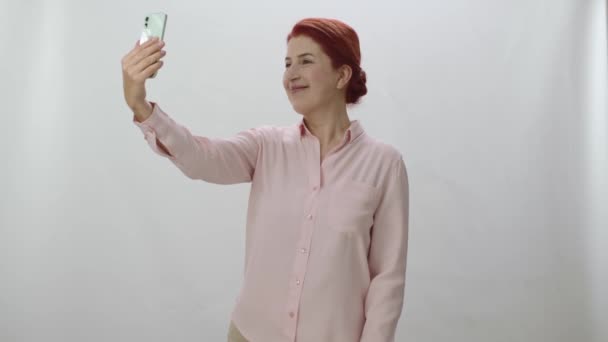白い背景に携帯電話で自撮りしている赤毛の女性 笑顔で携帯電話にポーズをとる幸せな女性 勝利サインを与えます — ストック動画