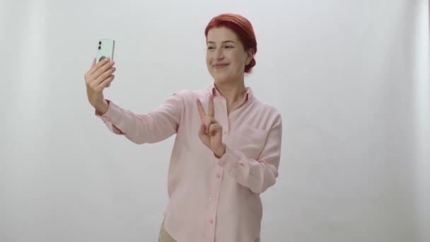 红头发女人拿着手机在白色背景下 快乐的女人面带微笑地在手机上摆姿势 给胜利的信号 — 图库视频影像