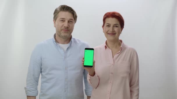 一对夫妇拿着一部带有绿色屏幕 白色背景的智能手机 男人指着智能手机用绿色屏幕做胜利的标志 你可以把你想要的图像放在绿色的屏幕上 — 图库视频影像
