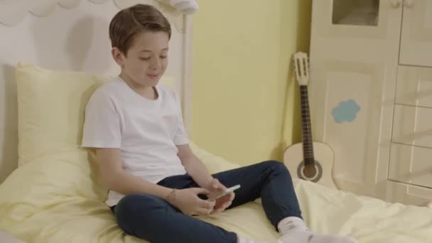 小さな男の子がスマートフォンを使ってベッドの上に座っている 子供は インターネットをサーフィンし 彼の携帯電話でビデオを見て オンラインゲームをしています 子供の技術利用の概念 — ストック動画
