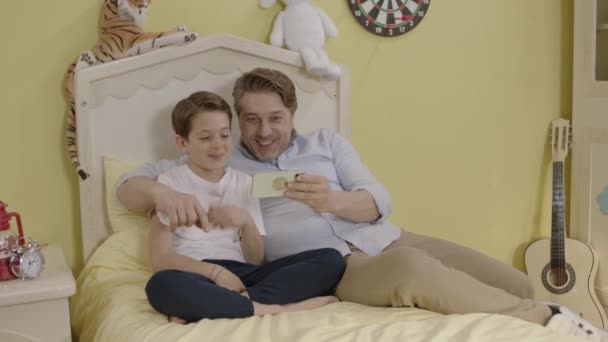 아버지가 스마트폰을 사용하고 침대에 아들을 미소짓는 아빠와 귀여운 침대에 집에서 — 비디오