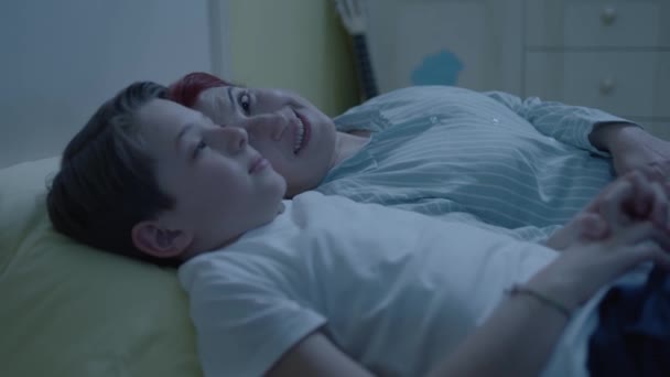 小さな息子とベッドの中に寝そべっている陽気な母親のイメージ 若い母親と男の子のベッドで 一緒に時間を過ごすチャット その少年は一日中何をしているのか母親に話す 理想的な母像 — ストック動画