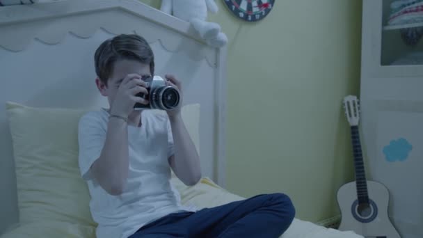 穿着睡衣坐在床上的漂亮男孩审视着一台旧相机 小男孩试图用他的相机拍照 聪明男孩对绘画感兴趣的画像 — 图库视频影像