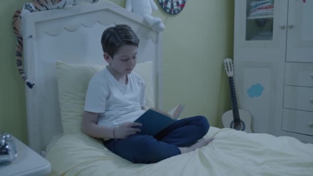 少年は夜寝室のベッドで本を読んでいる 家にいるパジャマ姿の男の子明るい笑顔の男の子が部屋のベッドの上に座って本を読んでいる — ストック動画