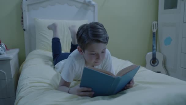 少年は夜寝室のベッドで本を読んでいる 子供は本を読む 家でパジャマを着た少年 彼の部屋のベッドの上に横になって本を読んでいます — ストック動画