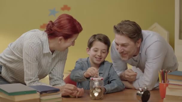 お金節約の概念 お母さん 小さな息子と夫は貯金箱にお金を入れている 幼い親は息子にお金を貯める方法を教える — ストック動画