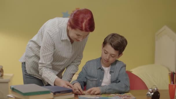 小さな男の子は 彼の机の上に母親とジグソーパズルをやっています 少年は余暇にパズルを解く 子供の世話をする理想的な母親の肖像画 — ストック動画
