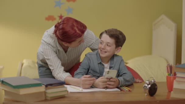幼い母親が子供がスマートフォンで宿題を聞くのを手伝っている 若い愛情のある母親は スマートフォンの使い方を彼女の小さな息子に教え 彼のオンラインレッスンを少年に示します 子供の発達の概念 — ストック動画