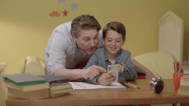 子供がスマートフォンで宿題を聞くのを手伝っている若い父親 幼い思いやりのある父親が息子にスマートフォンの使い方を教えており 少年にオンラインレッスンを見せています 子供の発達の概念 — ストック動画