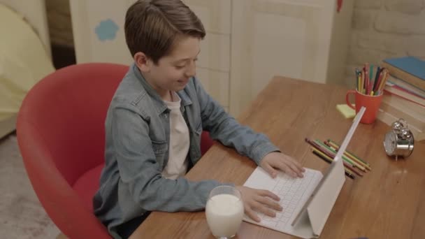 タブレットでレッスンをしながら小さな男の子が牛乳を飲んでいる 彼はコンピューターのレッスンを受け ゲームをしたり 部屋の机で牛乳を飲みながらビデオを見たりします ミルクは健康 — ストック動画