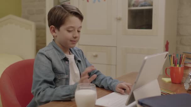 タブレットでレッスンをしながら小さな男の子が牛乳を飲んでいる 彼はコンピューターのレッスンを受け ゲームをしたり 部屋の机で牛乳を飲みながらビデオを見たりします ミルクは健康 — ストック動画