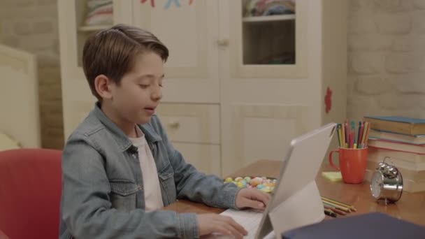 小男孩一边在平板电脑上上课 一边吃榛子 他一边在房间的桌子上吃色彩艳丽的糖果 一边上计算机课 玩游戏 看录像 — 图库视频影像