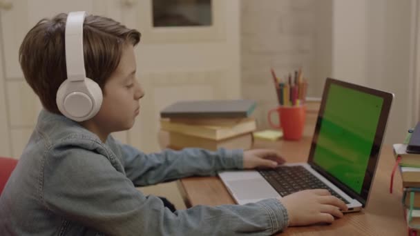 緑の画面のノートパソコンでヘッドフォンを使用して自宅からオンラインで働いている小さな学校の学生は ビデオ通話で教師に耳を傾けます 画像を緑の画面に配置できます — ストック動画