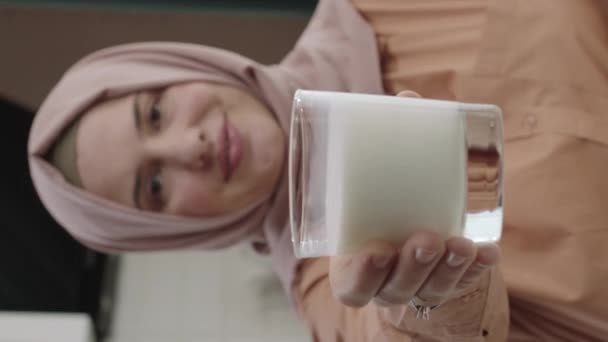 ヒジャーブ州の女性はカメラにミルクのガラスを与えます 健康的な飲み物の概念 台所のテーブルに座ってカメラにミルクを与えるヒジャーブの若い女性の笑顔 垂直ストーリーのビデオ — ストック動画