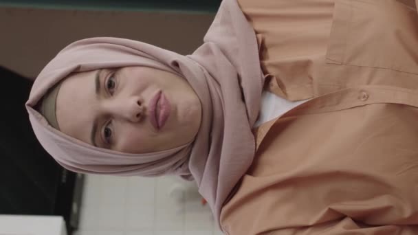キッチンに立ってカメラを見て服を着たヒジャーブの若い大人のイスラム教徒の女性の笑顔を自信を持って 若い大人の女性がカメラを見て微笑んだ ムスリム女性の肖像画 垂直ストーリーのためのビデオ — ストック動画