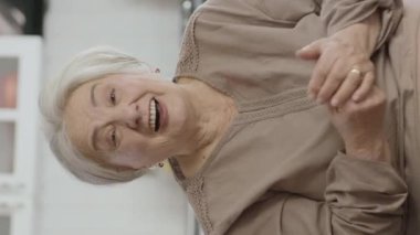 Gülümseyen yaşlı kadın video görüşmesi yapıyor. Kameraya el sallıyor. Dikey hikaye için ön kamera görüntüsü..