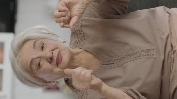 高齢の白髪の女がリビングルームのソファの上に薬を指す 彼女は彼女の手の薬が優れていることを指摘している 幸せな老女ショーVitamins Vertical Storyビデオ — ストック動画