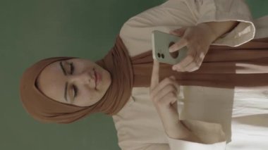 In the classroom,the teacher in hijab looks at social media accounts with her smartphone,browses the internet,reads e-mails,surprised by what she sees and showing her green screen phone to the camera.