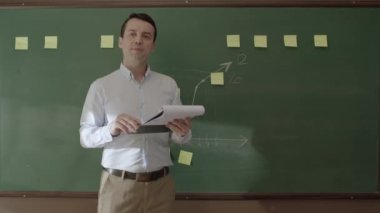 Male teacher looking at his notes in front of the blackboard, talking to his students. The teacher is reading reports in front of a green blackboard.