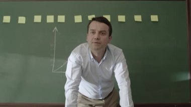 Male teacher standing in front of blackboard with glued notes and chalk drawn math formulas, smiling at camera and teaching his students. Online distance education.