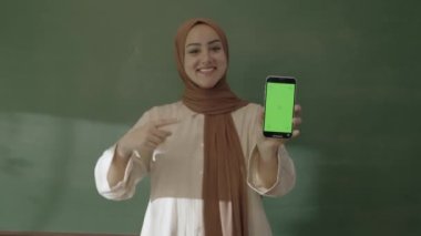 In the classroom,the teacher in hijab looks at social media accounts with her smartphone,browses the internet,reads e-mails,surprised by what she sees and showing her green screen phone to the camera.