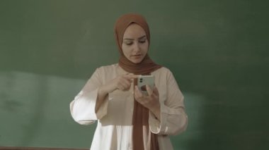 In the classroom,the teacher in hijab looks at social media accounts with her smartphone,browses the internet,reads e-mails,surprised by what she sees and showing her green screen phone to the camera.