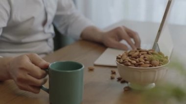 Businessman drinking coffee while working remotely from home. Freelance man drinking coffee, eating roasted almonds, hazelnuts, cookies while working remotely at computer.