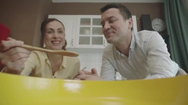 View of people cooking from a pot or pan. They are tasting the food in the cooking process with a wooden spoon in the kitchen. The camera is inside the pot.