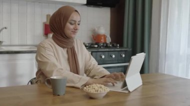 Young woman in hijab works remotely on a tablet computer. The woman drinks coffee or tea and eats roasted hazelnuts while working on the computer in her home kitchen. Freelancer concept.