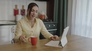 Young woman working remotely on tablet computer. The woman is drinking coffee or tea in a cup while doing her chores in the kitchen of her home. Freelance concept.