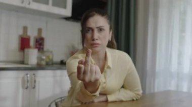 Young woman showing middle finger, angry and disobedient at bad behavior. In the kitchen, the middle finger points badly at the camera and asks not to be disturbed. View from computer camera.