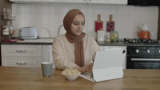 ヒジャーブの若い女性がタブレットコンピュータで遠隔操作してる 自宅のキッチンでパソコンを操作しながらローストヘーゼルナッツを食べていた フリーランスのコンセプト — ストック動画