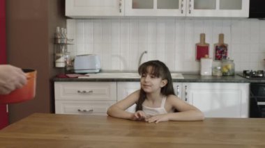 The little girl waits at the table for the meal prepared by her mother. When the girl lifts the lid of the pot, she is very happy to see that her mother is cooking her favorite dish.